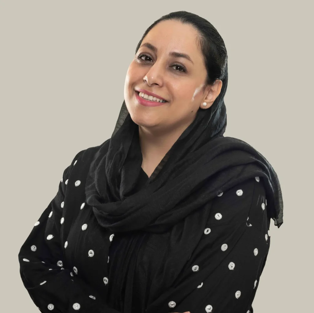 تصویر فاطمه حافظ نیا با مانتو مشکی توپ توپی سفید با شال مشکی دست به سینه و مایل به چپ ما ایستاده و داره لبخند میزنه